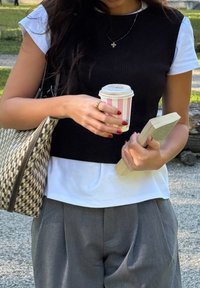 Crna rebrasta majica bez rukava preko bijele košulje, sivo plisirane hla�če, drži prugastu šolju za kafu i knjigu, uz šareni torbu.