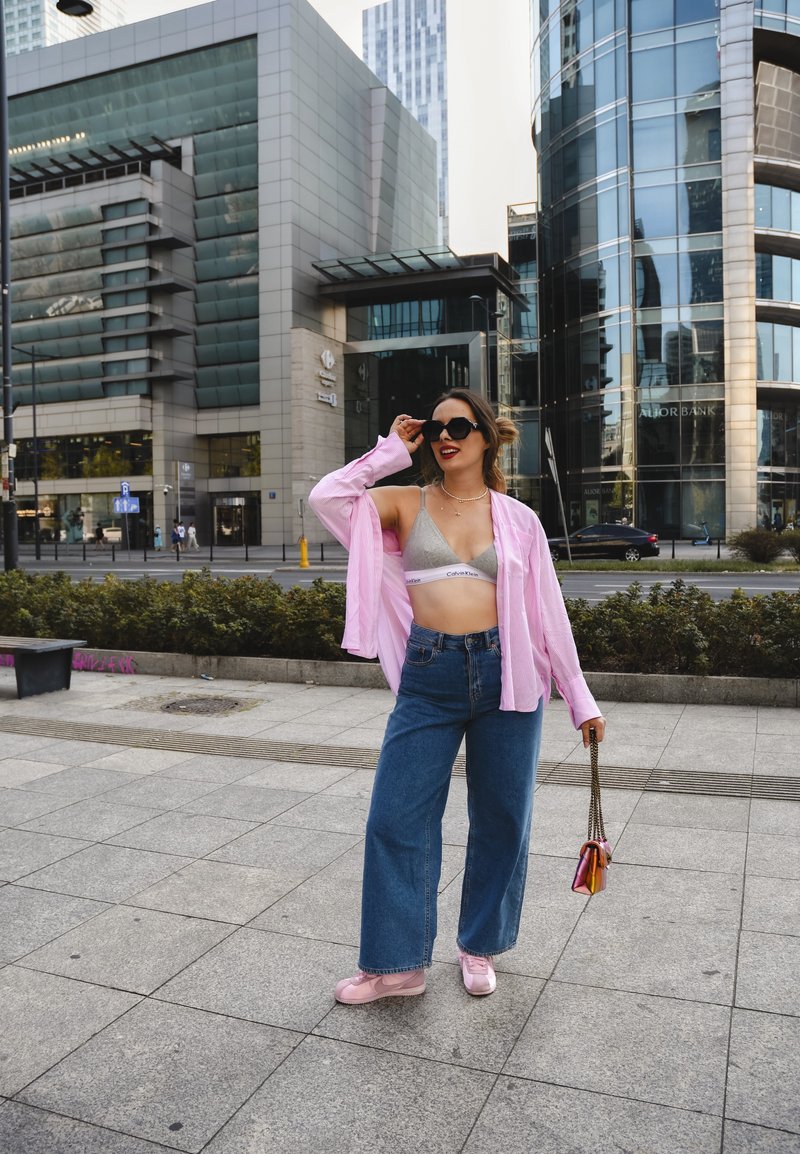 Φωτεινό ροζ υπερμεγέθη πουκάμισο πάνω από ένα γκρι μπράλετ, συνδυασμένο με φαρδιές μπλε τζην και ροζ αθλητικά παπούτσια. Κρατώντας μια πολυχρωμία τσάντα.