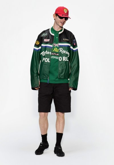 Chaqueta de moto de cuero verde con rayas azules y blancas, camiseta de polo verde, pantalones cortos negros, gorra roja y zapatillas deportivas negras.