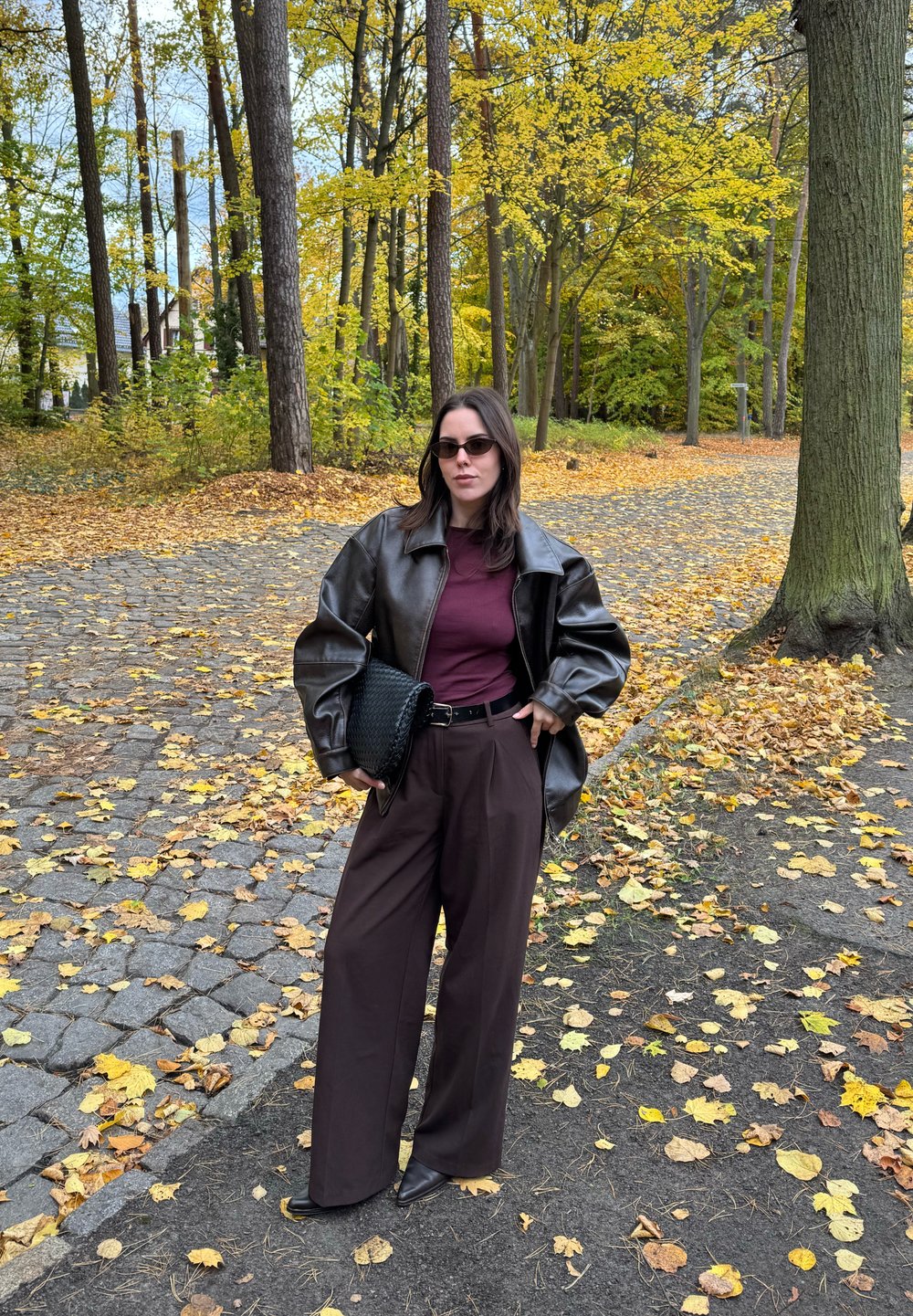 Mujer con gafas de sol, vistiendo una chaqueta de cuero marrón, una blusa burdeos y pantalones anchos marrones, sosteniendo un bolso negro, de pie en un camino cubierto de hojas en un bosque en otoño.