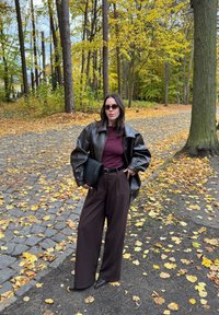 Mujer con gafas de sol, vistiendo una chaqueta de cuero marrón, una blusa burdeos y pantalones anchos marrones, sosteniendo un bolso negro, de pie en un camino cubierto de hojas en un bosque en otoño.