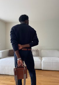 Person steht drinnen und hält eine braune Ledertasche hinter dem Rücken vor einem weißen Sofa mit Kissen.