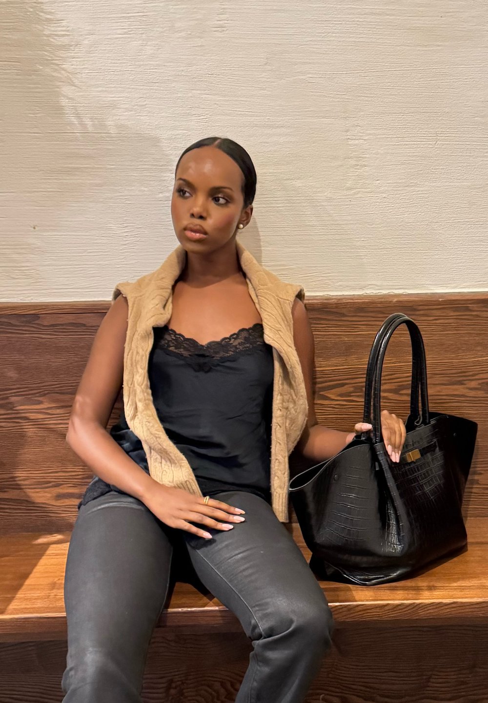 Une femme est assise sur un banc en bois, portant un haut en dentelle noire, un gilet sans manches beige, un pantalon gris, tenant un grand sac à main noir à côté d'elle sur le banc.
