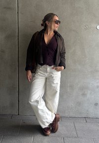 Giacca bomber marrone su top a coste prugna, abbinata a pantaloni bianchi a gamba larga. Scarpe con zeppa marroni presentano dettagli dorati e lacci.