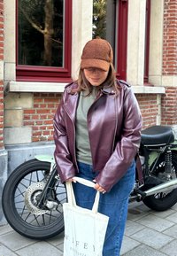 Bordeaux læderjakke med lynlås foran, gråt top og blå jeans, holder en cremefarvet tote taske. Baggrunden inkluderer en motorcykel og en murstensvæg.