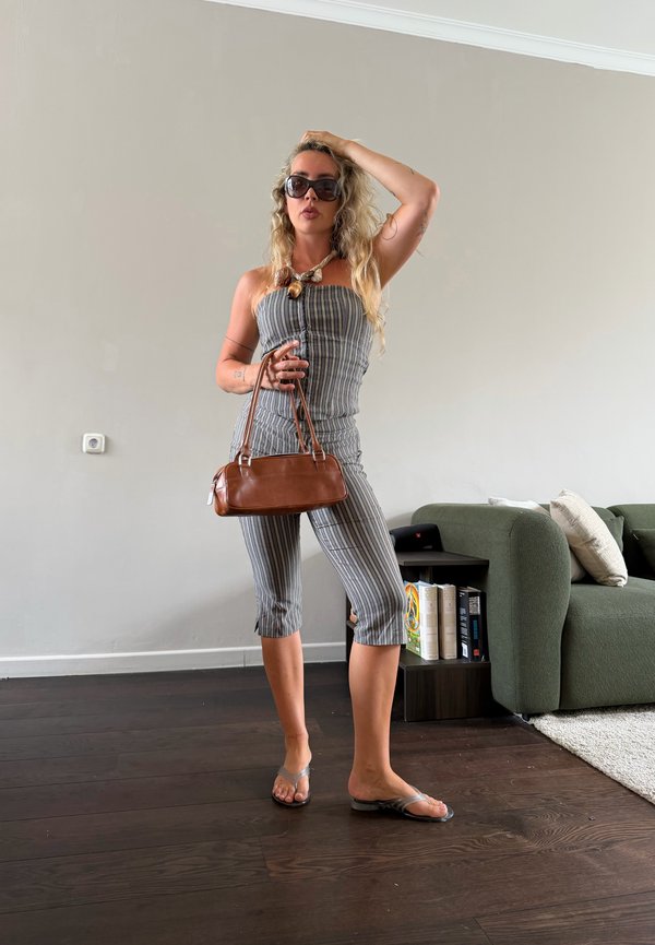 Frau in gestreiftem schulterfreiem Oberteil und Hose hält eine braune Handtasche, trägt große Sonnenbrille und Flip-Flops, steht im Wohnzimmer mit grünem Sofa und Bücherregal.