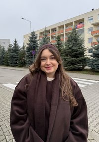 Bruine, oversized fleece jas met een sjaalkraag, gecombineerd met een licht truitje. Lang, golvend haar, tegen een achtergrond van groenblijvende bomen en gebouwen.