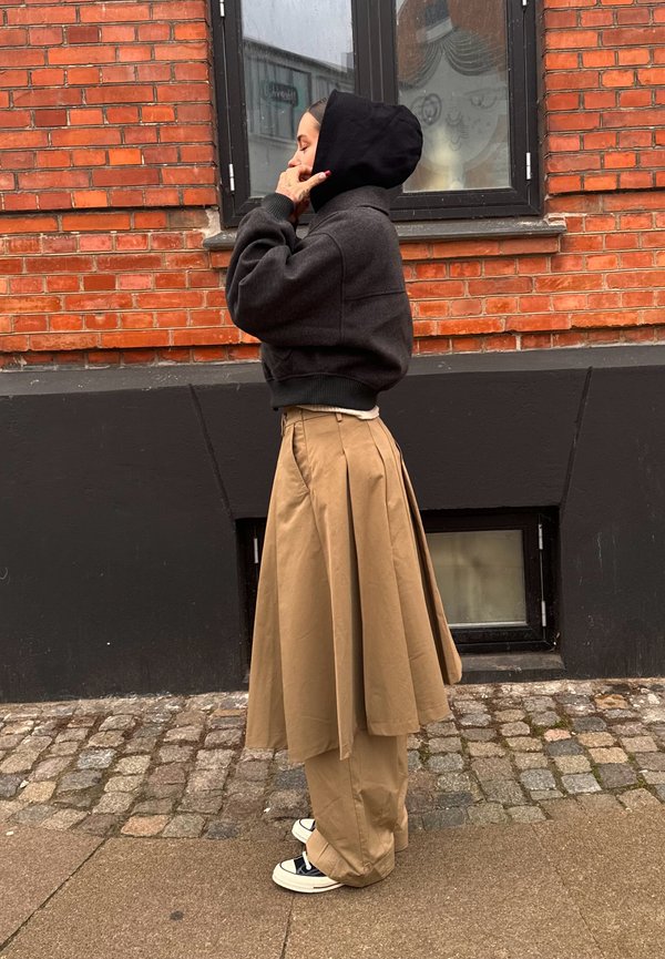 Hoodie cinza cortado com capuz preto, combinado com uma saia plissada bege. Usado sobre calças largas bege e ténis preto e branco. Fundo de parede de tijolo.