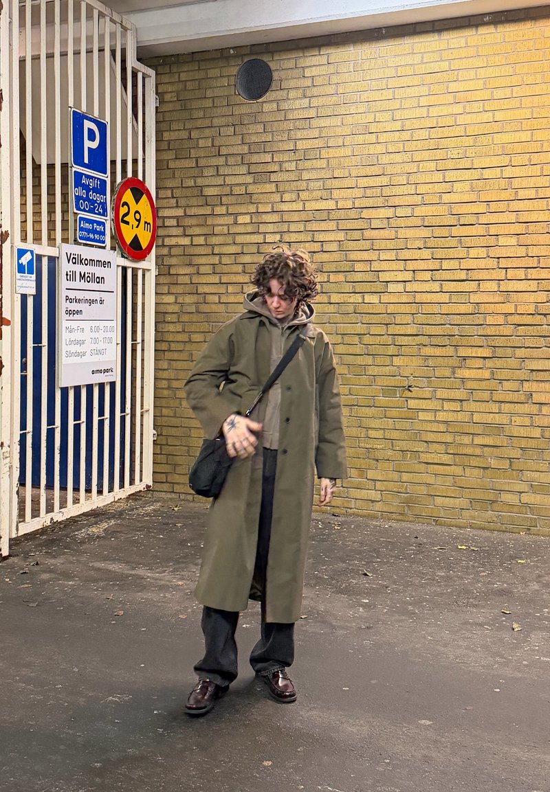 Cappotto lungo verde oliva sopra una felpa grigia, pantaloni neri e scarpe marroni. Borsa nera a tracolla, in piedi contro un muro di mattoni gialli.