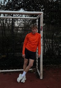 Odzież sportowa: pomarańczowa bluzka z długim rękawem i suwakiem, czarne spodenki, białe trampki. Ustawiona przy słupku bramki na tle sportowego otoczenia.