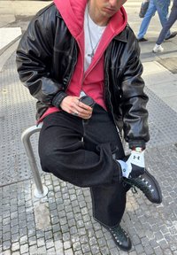 Jeune personne assise sur une barrière métallique de rue, portant une veste en cuir noire, un sweat à capuche rouge, un pantalon noir, des gants et tenant une tasse de café.