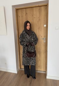 Manteau à motif léopard avec grand col, écharpe tricotée marron, gants foncés, pantalon noir et sac à main marron, debout près d'une porte en bois.