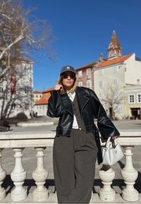 Jaqueta de couro preta, camisa branca, calças cinzas e um boné cinza. Óculos de sol e uma bolsa branca com detalhes metálicos. Cenário urbano.
