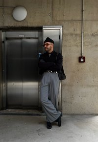 Άνδρας με σταυρωμένα τα χέρια, φορόντας μαύρο πουλόβερ και γκρι παντελόνι, στέκεται δίπλα σε μεταλλικό ασα νσέρ πάνω σε σκυρόδεμα τοίχο. Μαύρα παπούτσια και καπέλο.