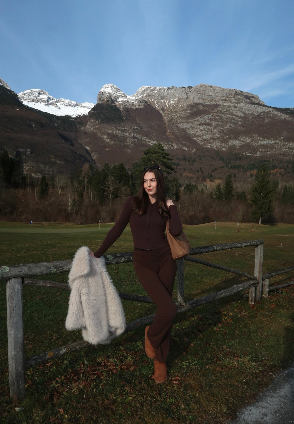 Conjunto marrón ajustado con una chaqueta con cremallera y pantalones acampanados, combinado con un abrigo crema y esponjoso colocado sobre una cerca de madera. Montañas nevadas al fondo.