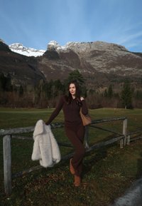 Conjunto marrón ajustado con una chaqueta con cremallera y pantalones acampanados, combinado con un abrigo crema y esponjoso colocado sobre una cerca de madera. Montañas nevadas al fondo.