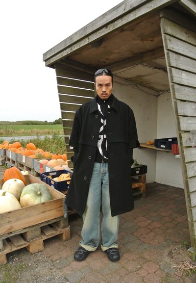 Abrigo negro con cuello, camisa a rayas debajo, jeans de mezclilla azul claro y zapatos negros. El fondo presenta cajas de madera con calabazas.