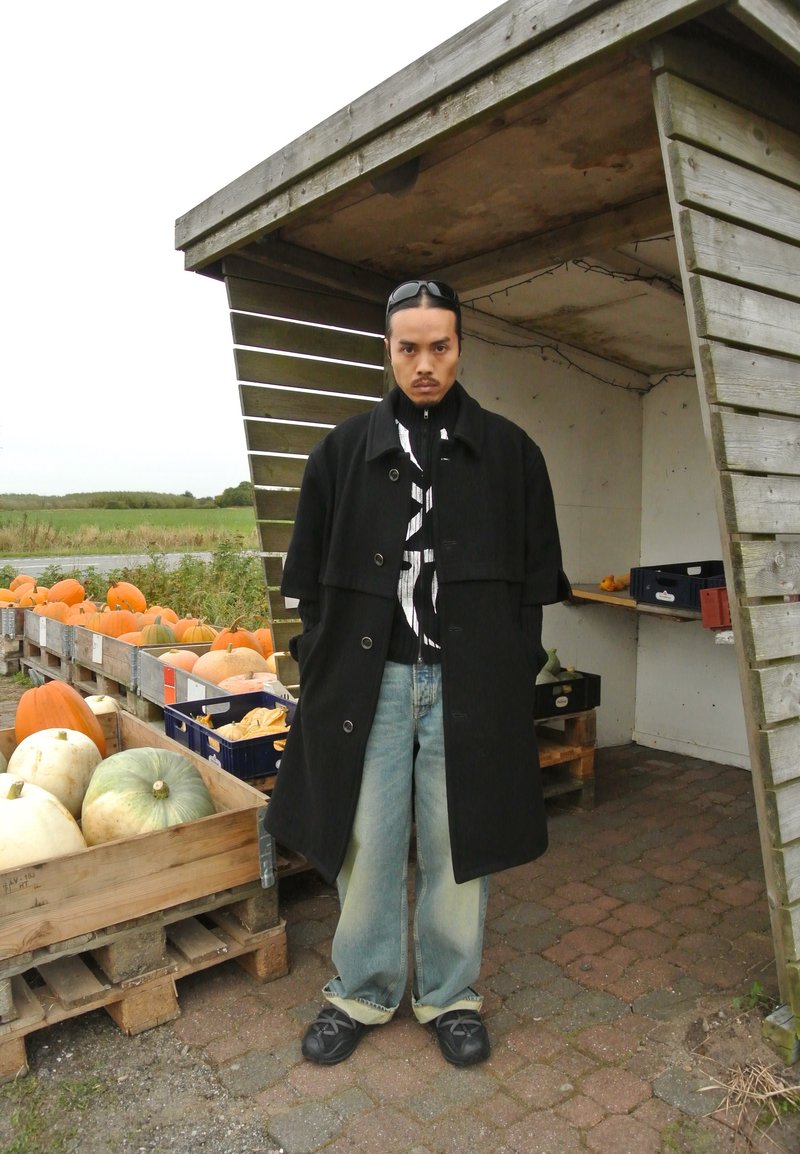 Μαύρο παλτό με γιακά, ριγέ πουκάμισο από κάτω, ανοιχτό μπλε τζιν και μαύρα παπούτσια. Το φόντο περιλαμβάνει ξύλινες κούτες με κολοκύθες.