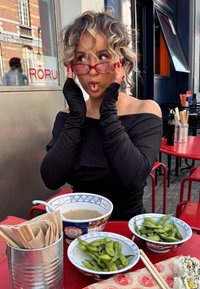 Svart off-the-shoulder topp, røde briller, krøllete hår. Skåler med grønn edamame og suppe på et rødt bord, med bestikk i en metallholder.