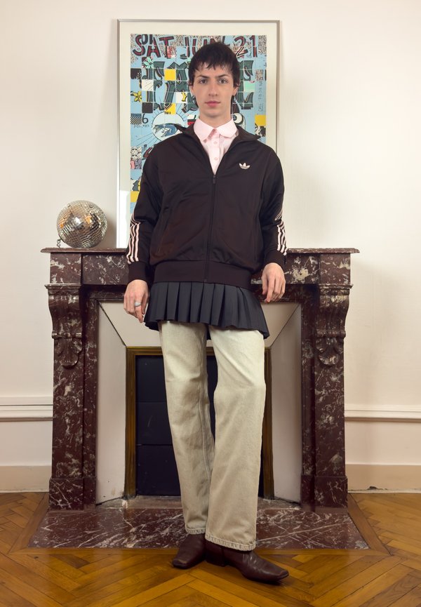 Ung person kledd i svart Adidas-jakke, plissert skjørt over beige bukser og brune støvler, står foran en marmorpeis med fargerik kunst.