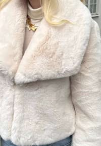 Kremhvit jakke i kunstpels med vid krage, myk tekstur og lukking foran. Brukes over en lys høyhalset genser, med et synlig gullkjede.