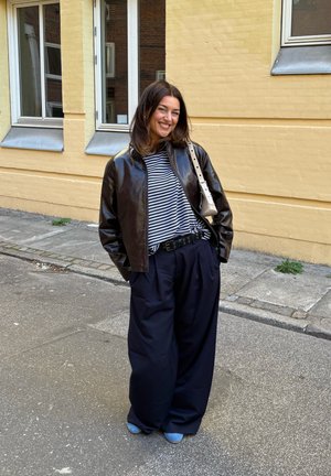 Chaqueta bomber de cuero marrón, pantalones de pierna ancha azul marino, camiseta a rayas, cinturón negro y zapatos azules. El fondo presenta una pared amarilla y ventanas.