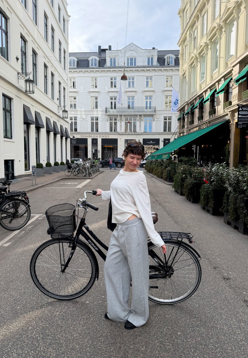 Personne en pull blanc et pantalon gris se tient à côté d'un vélo noir dans une rue de la ville avec des bâtiments historiques et des terrasses en plein air.