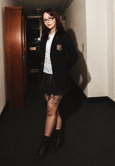 Joven mujer con cabello rojo y gafas que lleva una blazer negra, camisa blanca, falda plisada, piernas tatuadas y botas oscuras en un pasillo oscuro.