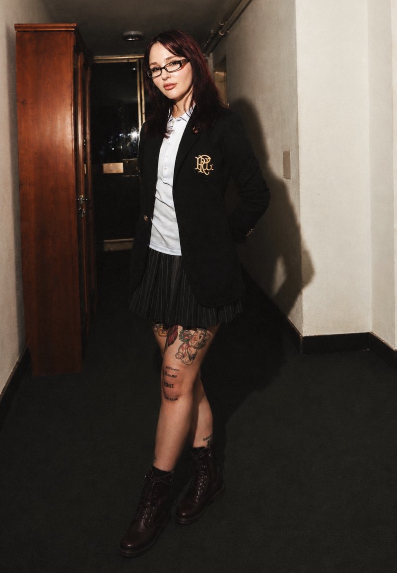 Junge Frau mit roten Haaren und Brille, die einen schwarzen Blazer, ein weißes Hemd, einen plissierten Rock, tätowierte Beine und dunkle Stiefel in einem dunklen Gang trägt.