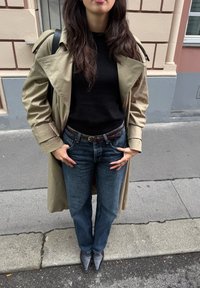 Béžový trench coat přes černý svetr, spárovaný s modrými džíny a špičatými černými kotníkovými botami. Textura: hladký kabát, denimová tkanina.