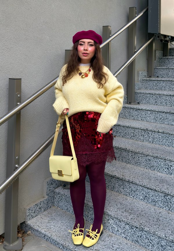 Femme portant un pull jaune, une jupe rouge à sequins, des collants bordeaux, des chaussures et un sac jaunes, ainsi qu'un béret magenta, debout sur un escalier près d'une rampe.