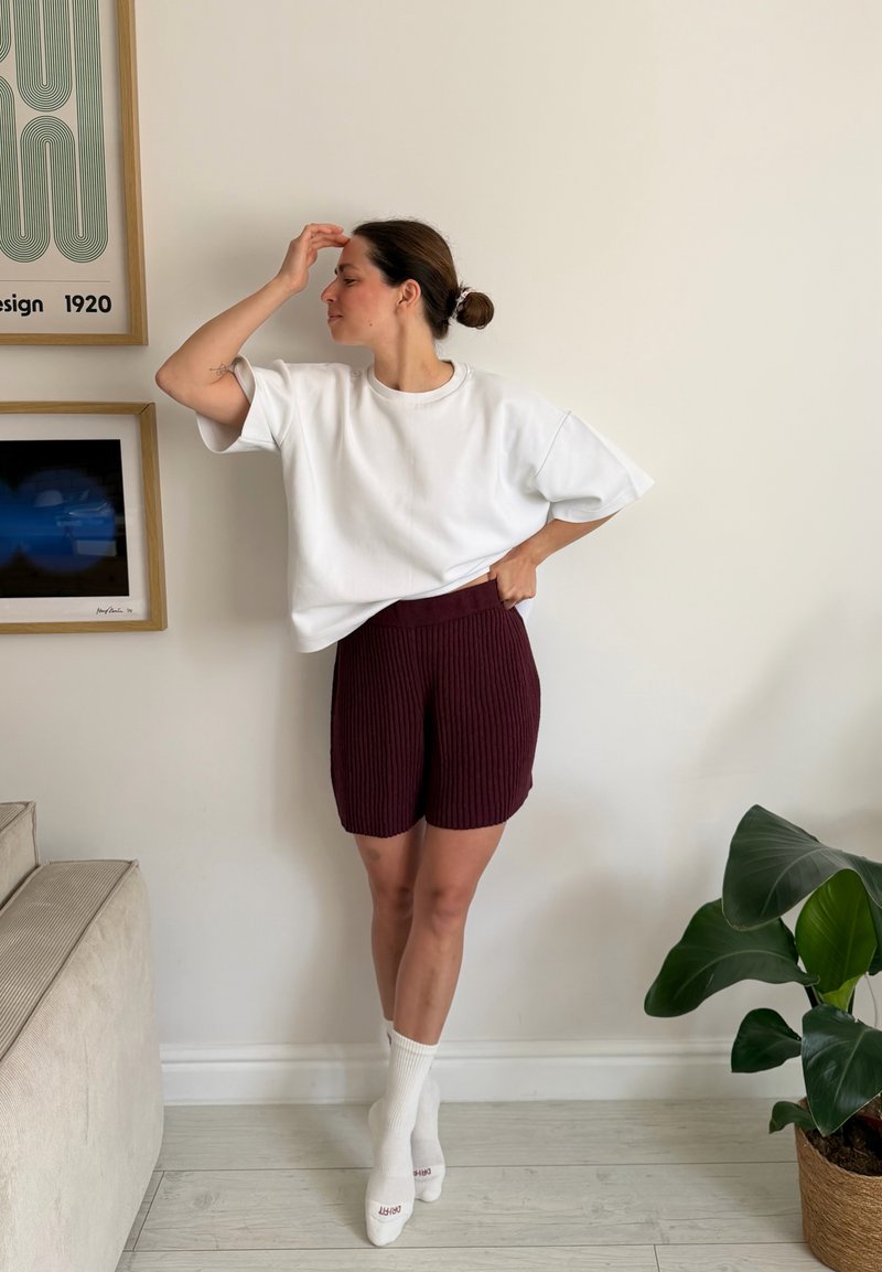 Kvinde, der står indendørs med håret sat op i en knold, iført en hvid oversized T-shirt, vinrøde ribstrikkede shorts og hvide sokker, der rører ved sin pande.