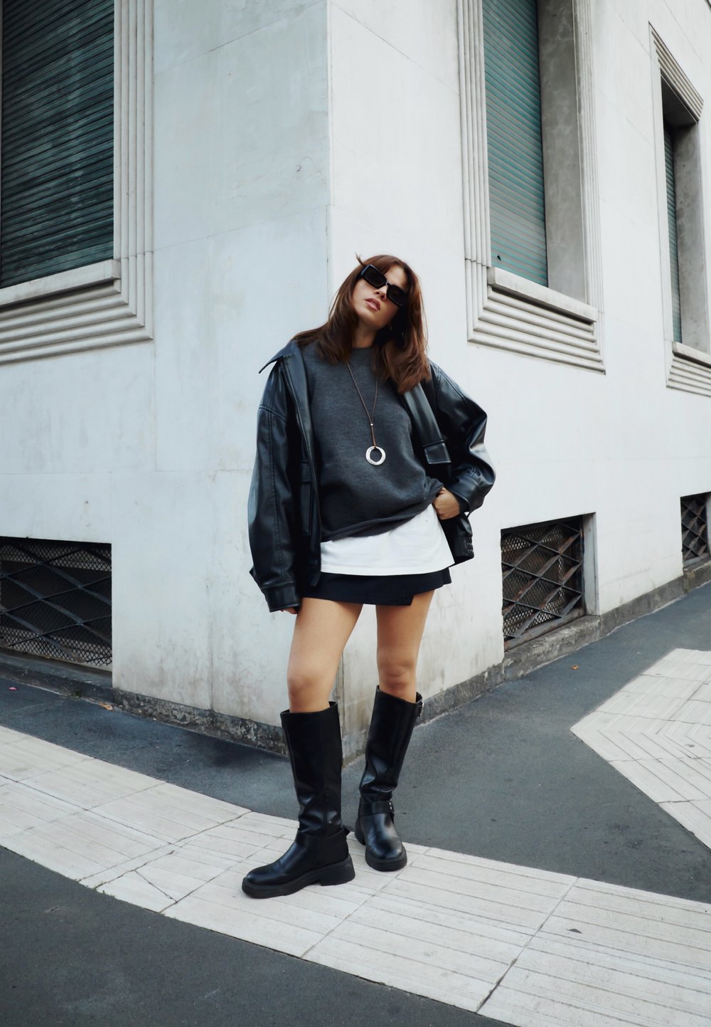 Čierna kožená oversize bunda, šedá mikina, biela vrstvená košeľa, čierna minibundička, čierne čižmy po kolená a veľké slnečné okuliare.