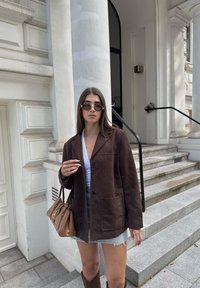 Chaqueta de ante marrón con cuello de pico, dos bolsillos frontales y cierre de botón, combinada con una blusa blanca y un bolso beige.