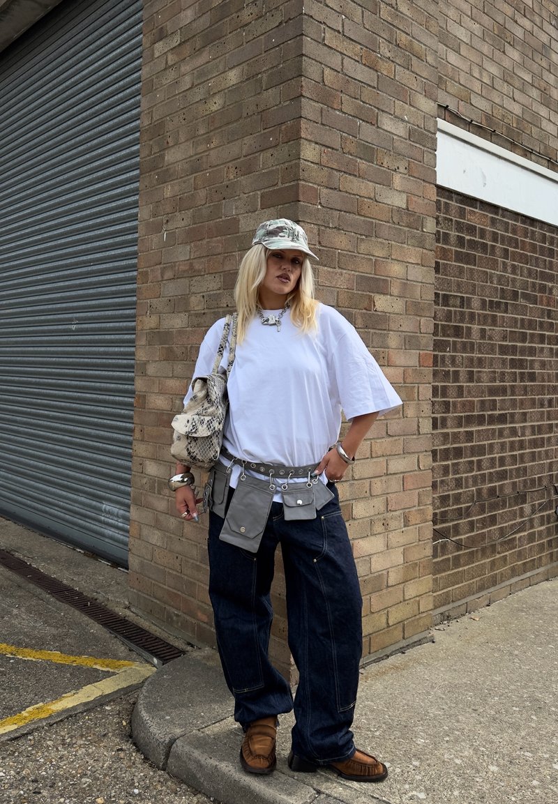 Femme portant un t-shirt blanc oversize, un jean ample foncé, des chaussures marron, une casquette camouflage et plusieurs petites pochettes grises sur une ceinture, debout près d'un mur en briques.