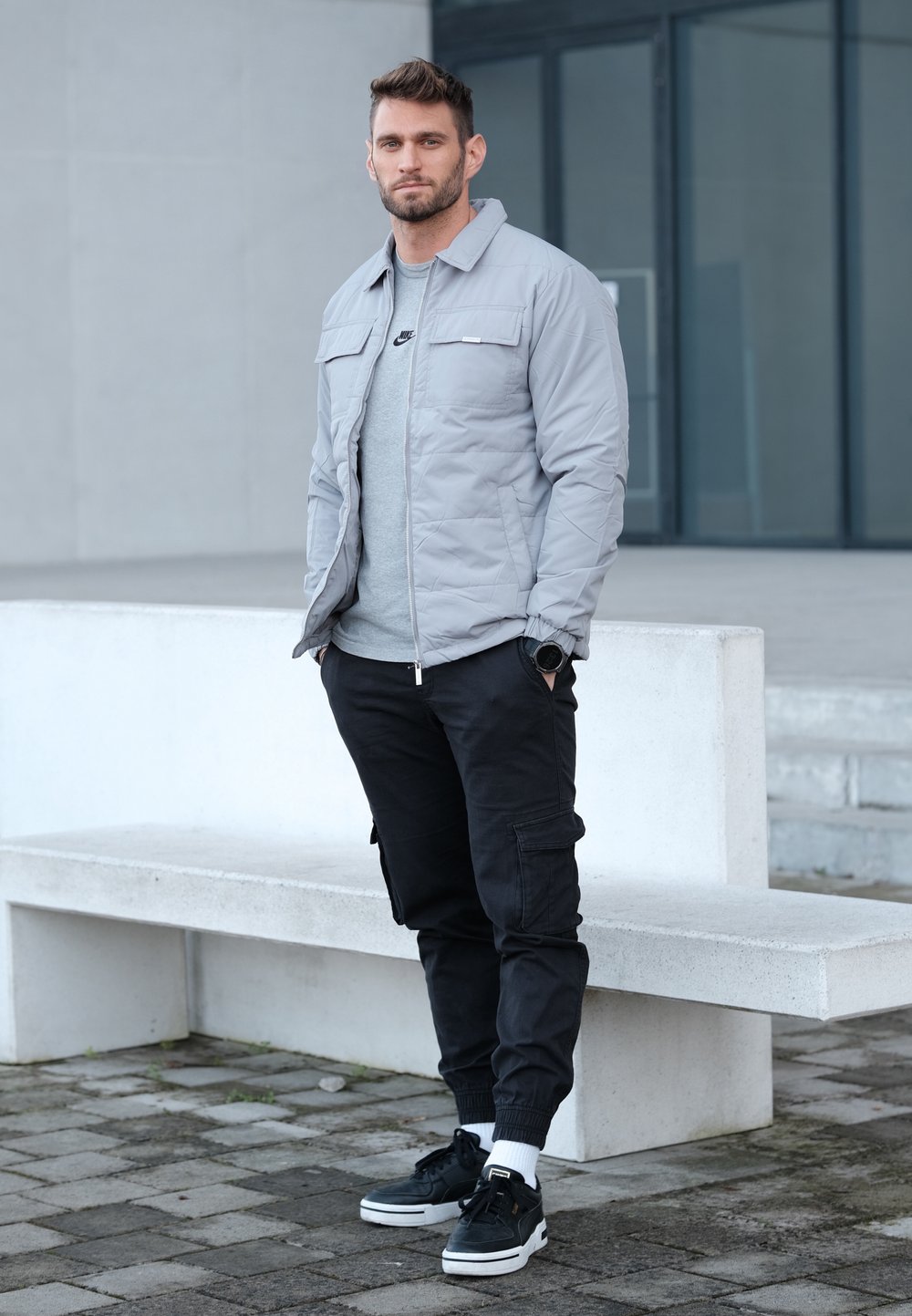 Veste matelassée gris clair portée sur un t-shirt gris, associée à un pantalon cargo noir et des baskets noires avec semelles blanches et chevilles resserrées.