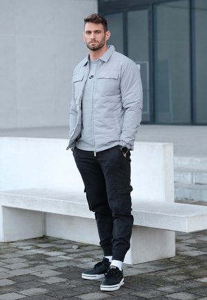 Chaqueta acolchada gris claro sobre una camiseta gris, combinada con pantalones cargo negros y zapatillas negras, con suelas blancas y puños en los tobillos.