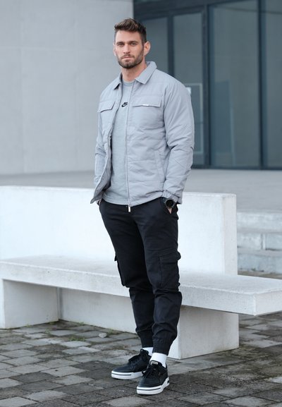 Chaqueta acolchada gris claro sobre una camiseta gris, combinada con pantalones cargo negros y zapatillas negras, con suelas blancas y puños en los tobillos.