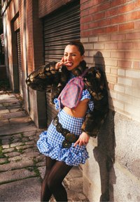 Femme portant une veste foncée en fausse fourrure, une robe à volants à carreaux bleus, des collants résille noirs et un sac rose à motifs. Elle porte une grande ceinture décorative.