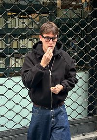 Ung mann i svart hettejakke og briller som påfører leppepomade mens han står foran en metallgitterport med stabler av bøker bak seg.