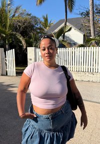 Kvinde iført en tætsiddende lyserød crop top og en lagdelt denimnederdel, som holder en sort taske. Baggrunden har palmer og et hvidt hegn.