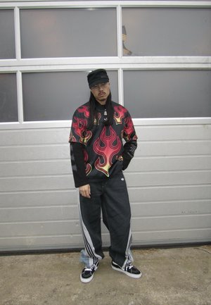 Hombre de pie frente a una puerta de garaje gris, lleva gafas, gorra negra, camisa con estampado de llamas sobre mangas largas, pantalones negros holgados y zapatillas deportivas.