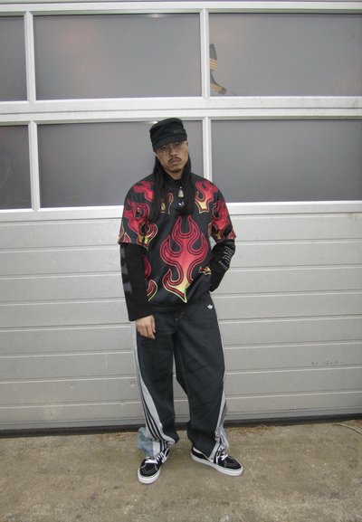 Hombre de pie frente a una puerta de garaje gris, lleva gafas, gorra negra, camisa con estampado de llamas sobre mangas largas, pantalones negros holgados y zapatillas deportivas.