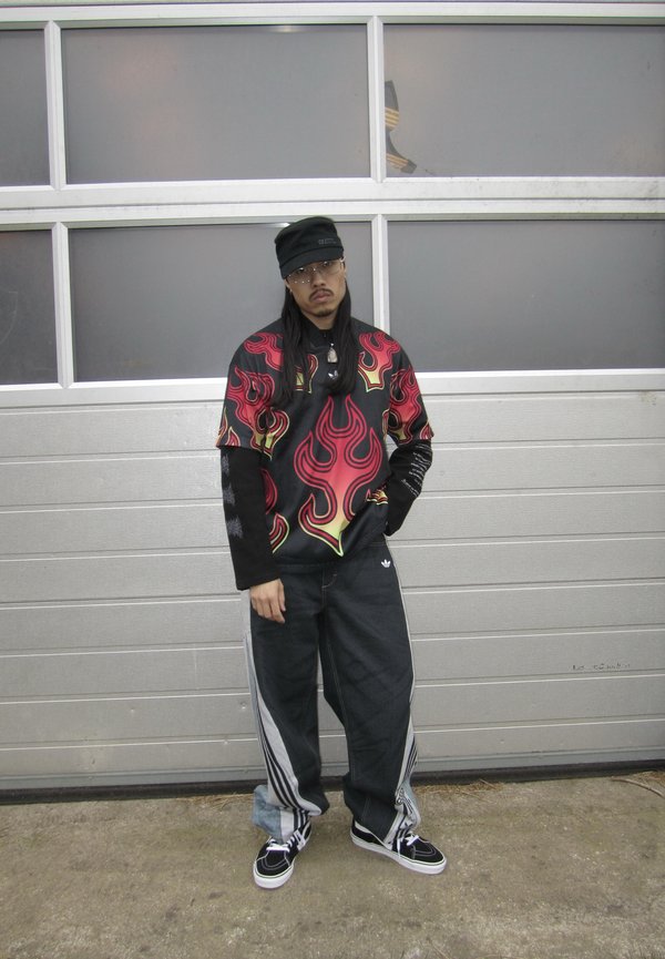 Mężczyzna stoi przy szarej bramie garażowej, nosząc okulary, czarną czapkę, koszulę z płomieniami na długich rękawach, luźne czarne spodnie i tenisówki.