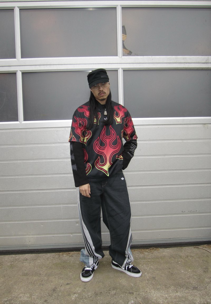 Homme debout devant une porte de garage grise, portant des lunettes, une casquette noire, une chemise avec motif de flammes par-dessus des manches longues, un pantalon noir ample et des baskets.