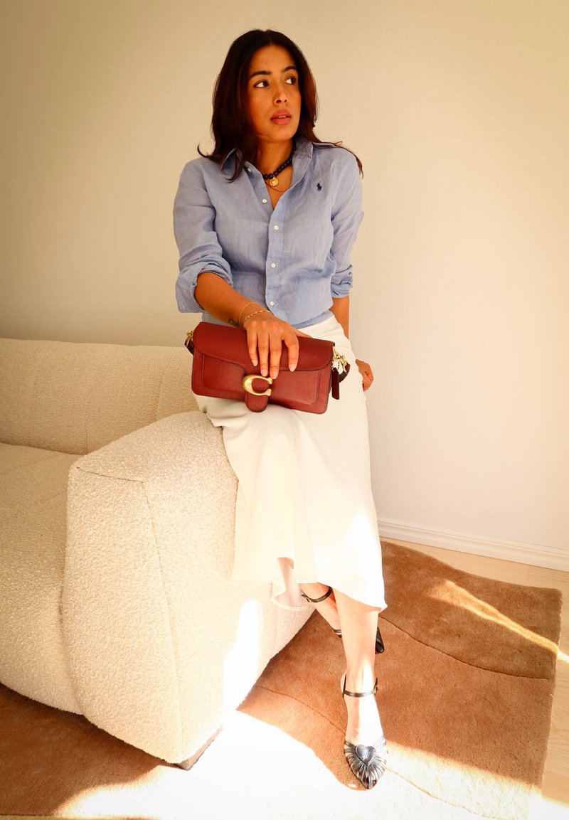 Woman in light blue shirt and white skirt sitting on beige sofa holding brown leather clutch bag on sunny carpeted floor.