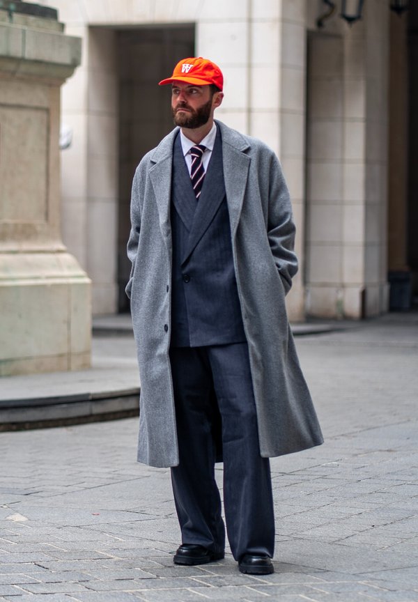 Un hombre barbudo con una gorra roja, un abrigo gris y un traje oscuro está de pie en una calle de la ciudad, junto a un edificio de piedra.