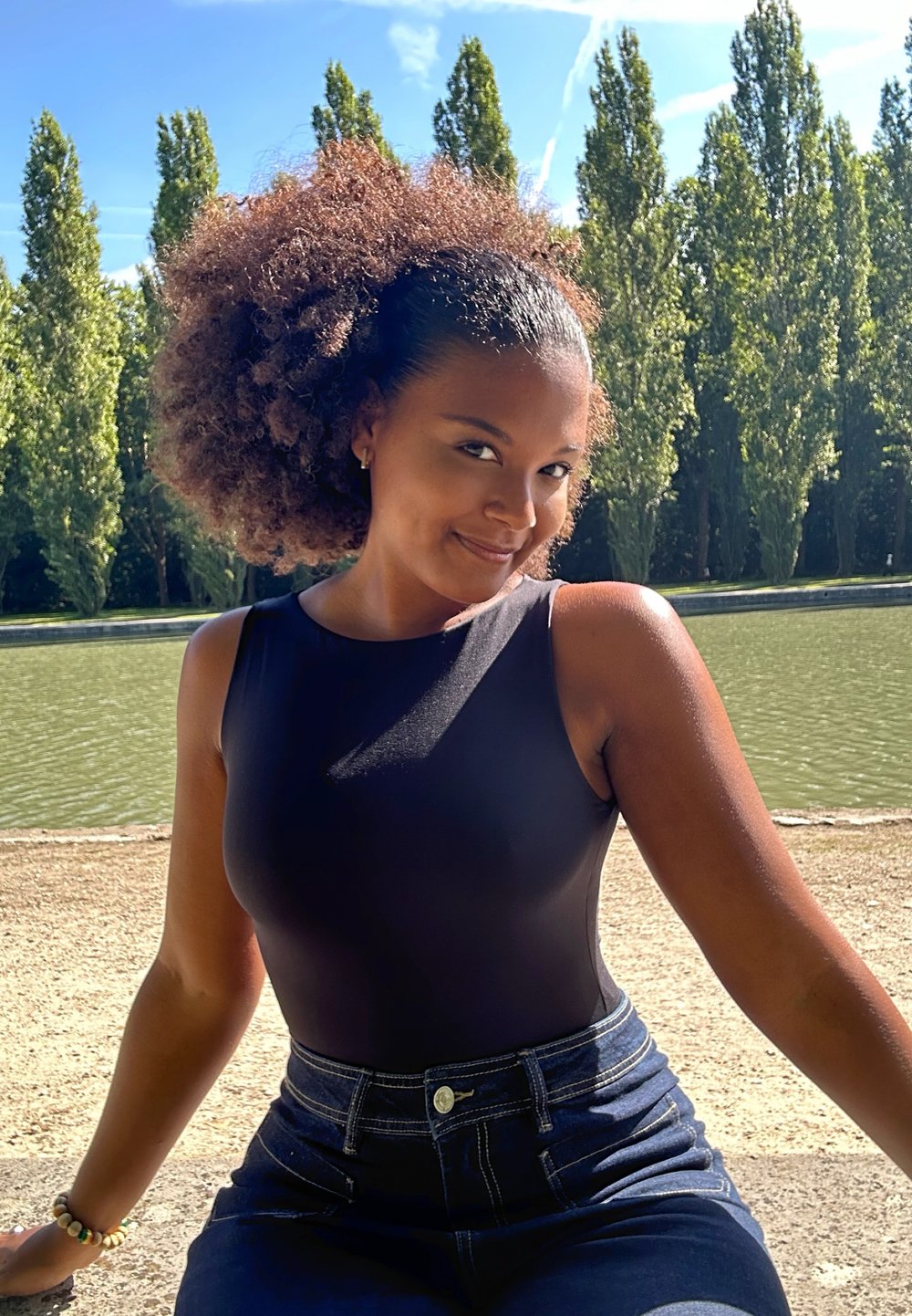 Schwarzer ärmelloser Bodysuit kombiniert mit hoch taillierten blauen Jeans. Lockige, voluminöse Haare; natürliche Außenumgebung an einem Wassermerkmal.