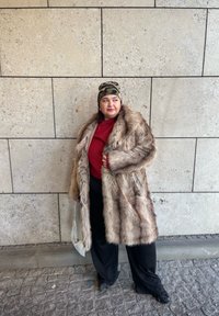 Manteau en fausse fourrure beige avec manches longues, associé à un pull rouge, un pantalon noir et un bonnet camouflage. Accessoire : un sac à main métallique.