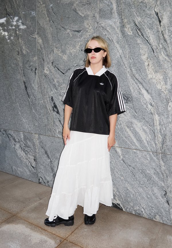 Donna con maglia Adidas nera, gonna bianca fluente, scarpe nere e occhiali da sole neri, in piedi contro un muro di marmo grigio.